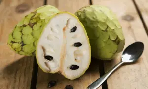 Dwa całe i przekrojone na pół cherimoya owoc z czarnymi pestkami, obok łyżeczka na drewnianym tle.