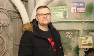 Mężczyzna w okularach i czarnej kurtce z futrzanym kapturem stoi na tle przemysłowych maszyn.