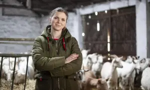 Kobieta w zielonej kurtce stoi na tle stada kóz. Jej uśmiech sugeruje, że hodowla kóz opłacalność jest dla niej satysfakcjonująca.