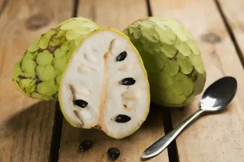 Dwa całe i przekrojone na pół cherimoya owoc z czarnymi pestkami, obok łyżeczka na drewnianym tle.