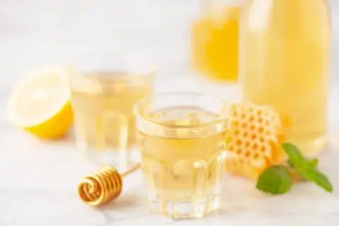 Złocista nalewka z jajek, cytryn, miodu i koniaku w kieliszkach, obok plastra miodu i cytryny.