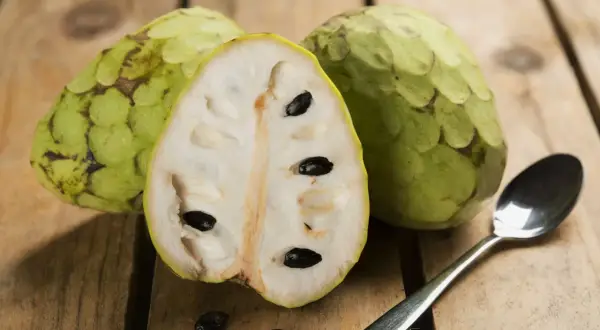 Dwa całe i przekrojone na pół cherimoya owoc z czarnymi pestkami, obok łyżeczka na drewnianym tle.