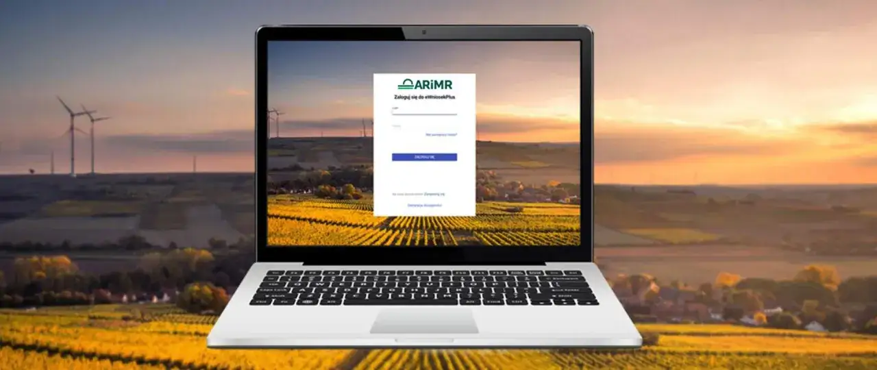 Laptop z widokiem na pola i wiatraki, na ekranie logowanie do systemu ARiMR. Sprawdź, jak sprawdzić wysokość dopłat bezpośrednich.