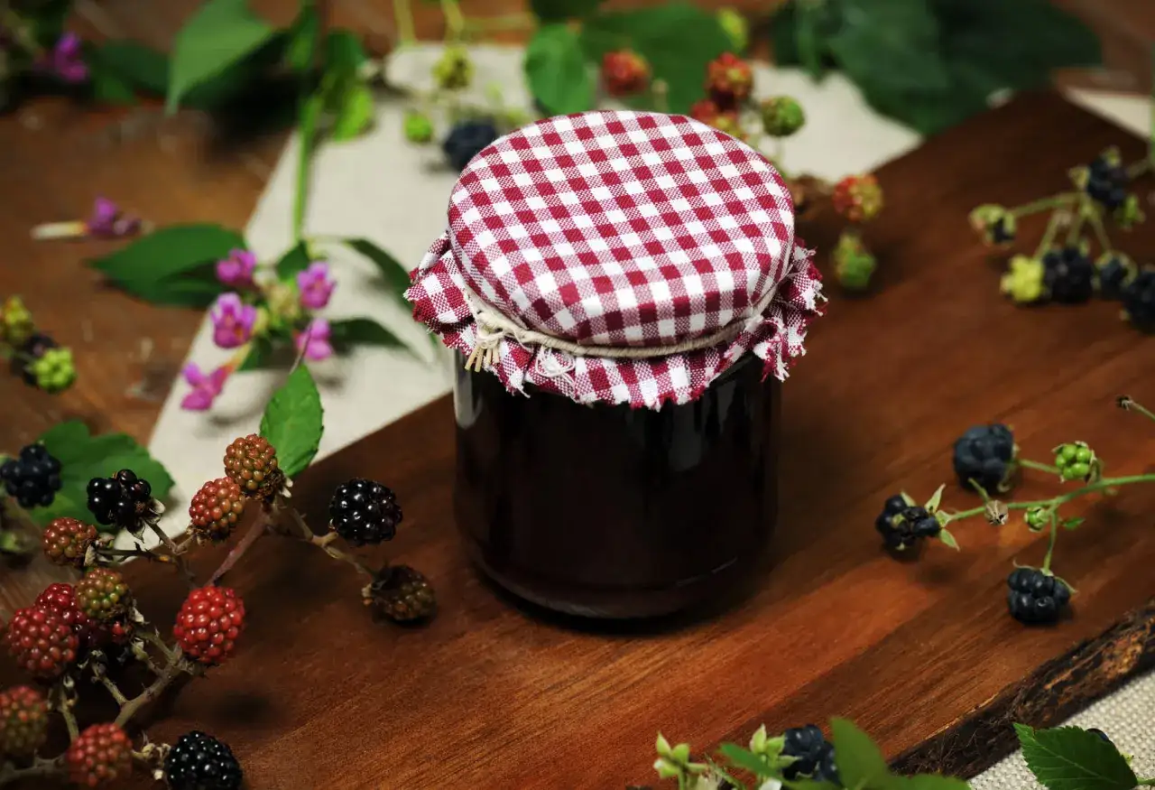 Słoik domowego dżemu z jeżyn, idealny na zimowe wieczory. Wokół świeże owoce i kwiaty. Co można zrobić z jeżyn? Dżem, nalewkę, a nawet ciasto!