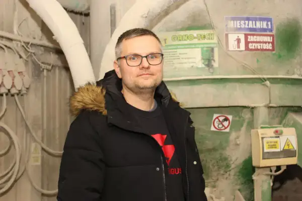 Mężczyzna w okularach i czarnej kurtce z futrzanym kapturem stoi na tle przemysłowych maszyn.